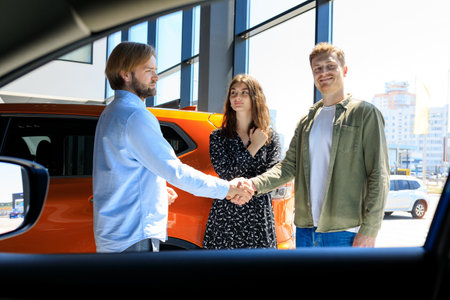 An employee of the dealership congratulates a married couple on buying a new car. Men shake hands.