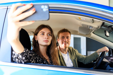 A young woman takes a selfie in the cabin of a new car against the background of a happy husband. Buying a car at a dealership.の写真素材