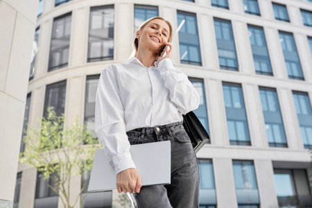 A well-groomed blond woman in a white blouse and black jeans stands against the background of an office building before getting a job.の写真素材