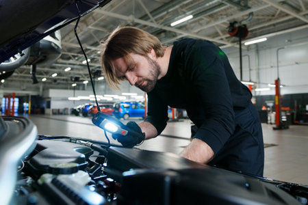 A male mechanic in uniform with a flashlight performs diagnostics under the hood of the car. Modern and technological car serviceの写真素材