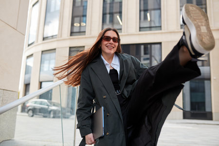 Stylish confident young freelance woman dressed in a black raincoat over a blouse walks near the...
