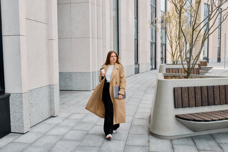 Business woman walks through the bustling city streets, exuding elegance in her trench coat, while clutching a laptop and sipping on a cup of coffeeの写真素材