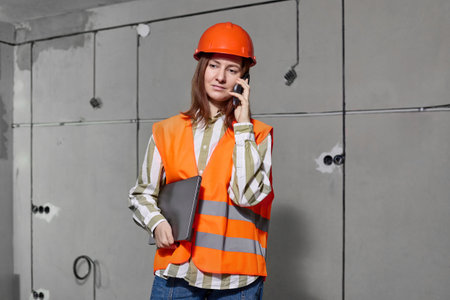 Young engineer builder woman in an orange vest and hard hat checks the correctness of electrical...