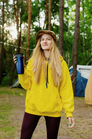 Camping in nature. Portrait of a tourist with a mug of tea against the background of a forest and tents. Vacation away from the cityの写真素材