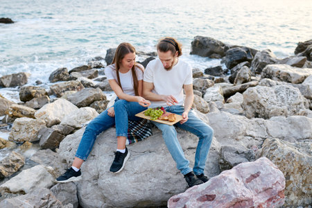 A man and woman share a meal on the rocky shore beside calm sea waters in evening light.の写真素材