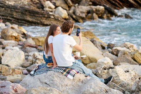 A man and a woman sitting on a rock, capturing a moment with a camera.の写真素材