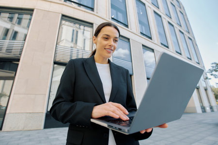 Pretty young business lady dressed in jacket works while walking around the city using laptop.の写真素材