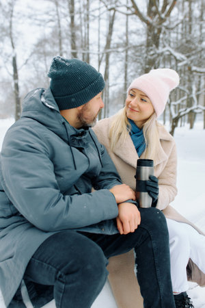 Couple enjoying a winter day in a snowy park while sipping hot beverages and sharing smiles.の写真素材