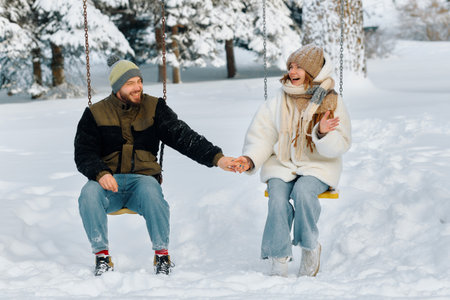 Couple enjoying a winter day on swings in a snowy park while holding hands and sharing laughter.の写真素材