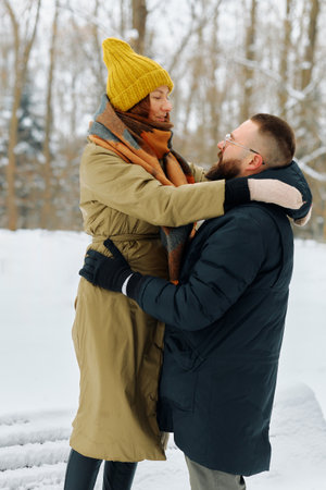 A couple stands in a snowy park, wrapped in warm clothing. The tall woman in a yellow beanie embraces her partner, creating a moment of happiness amidst the winter scenery.の写真素材