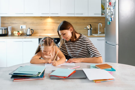 A mother helps her young daughter with homework at a kitchen table filled with supplies.