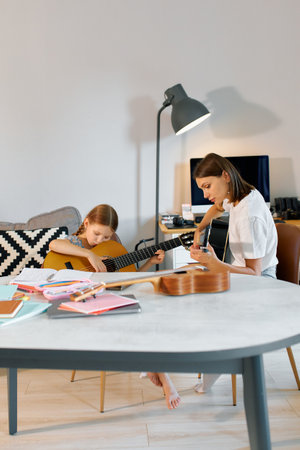 A mother teaches her daughter guitar in a stylish living room filled with sunlight.の写真素材