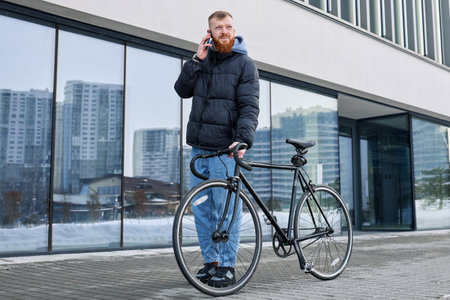 Red-haired Caucasian man in autumn clothes waiting for friend standing with bicycle near building.の写真素材