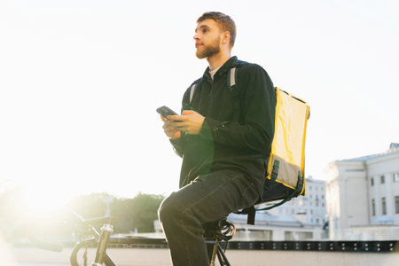 The bike courier looks at the delivery application in the phone. A man with a yellow backpack with a smartphone in his hands against the background of the city streetsの写真素材