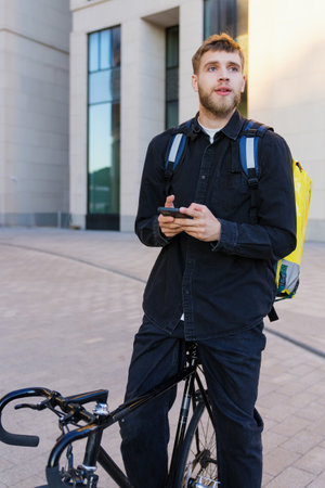 Vertical photo of a courier in a black shirt on a bicycle who is waiting for a new order. Using a smartphone at workの写真素材
