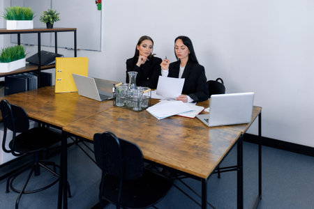 Two professional women engage in a detailed discussion at a large wooden table in a contemporary office space.の写真素材