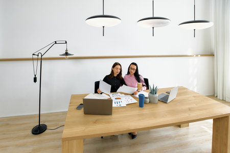Two women are engaged in a collaborative work session in a modern office, reviewing documents and discussing ideas at a large wooden table with laptops.の写真素材