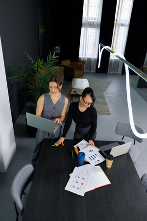 Two women work together in a stylish office setting during the day. One is holding a laptop while the other review documents on the table.の写真素材