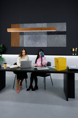 Two women are engaged in a discussion at a contemporary office setting, seated at a stylish black table with laptops and documents.の写真素材