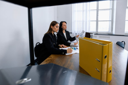 Two women in professional attire are sitting at a wooden table in a meeting room. They are reviewing documents.の写真素材