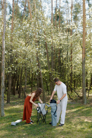 Couple prepares their camping chairs in a lush green forest. Sunlight filters through the leaves, creating a serene atmosphere for relaxation.の写真素材