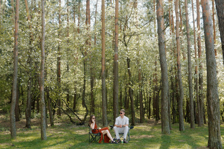 In a serene forest, a couple sits on folding chairs, enjoying each other's company amid blooming trees. The atmosphere is peaceful.の写真素材