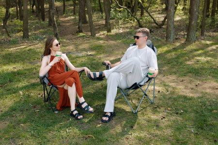 Two friends sit comfortably in folding chairs enjoying drinks in a serene park. Surrounded by trees, they share smiles and conversation.の写真素材