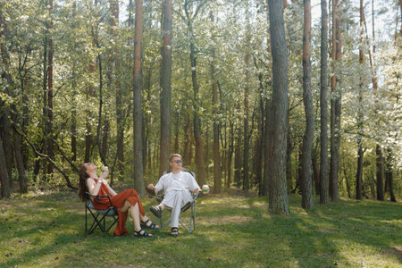Two individuals sit comfortably in folding chairs surrounded by tall trees and vibrant greenery. They enjoy cold drinks.の写真素材