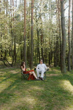 Two people enjoy a serene moment in the forest. They sit in folding chairs, surrounded by tall trees.の写真素材