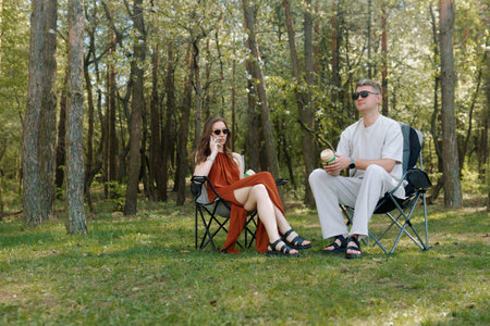 Two young adults sit comfortably in camping chairs at a park on a sunny day. The woman wears a flowing dress while the man wears sunglasses.の写真素材