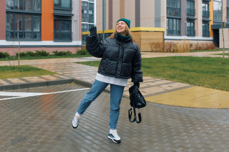 Woman waving cheerfully in a stylish winter outfit on a sidewalk outside modern buildings.の写真素材