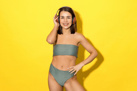 A laughing young woman in a swimsuit on a yellow background listens to music with headphones. Mood from a summer vacationの写真素材