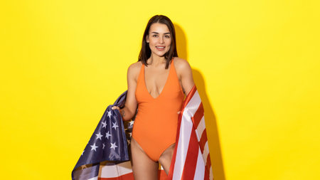 Bring a touch of patriotism to your summer holiday with this stunning photo featuring a young woman in a swimsuit proudly holding the American flag against a bright yellow backdropの写真素材