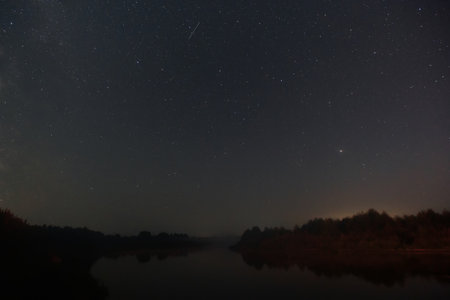 Under a vast night sky, countless stars twinkle above a tranquil river reflecting the celestial...
