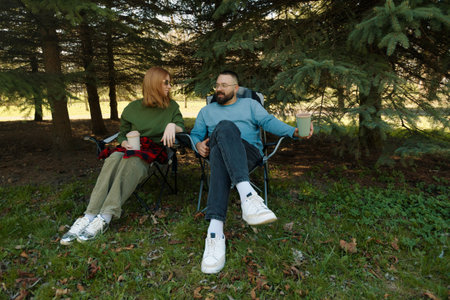A couple sits in folding chairs surrounded by trees, engaging in conversation and sipping drinks on...