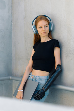 Woman with a bionic arm wears headphones and holds a laptop while leaning against a wall.