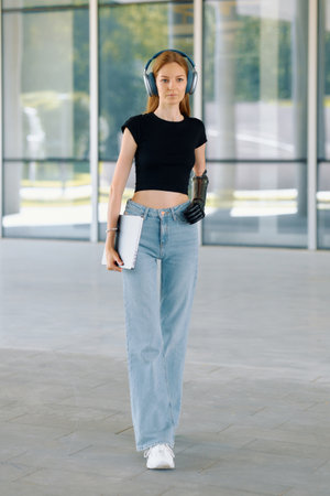 A woman with a bionic arm is walking outdoors, carrying a notebook and wearing headphones.の写真素材