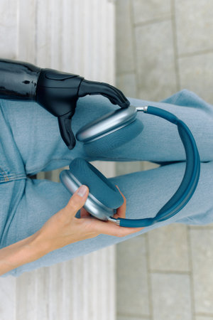 A woman with a bionic hand is holding a pair of headphones while sitting outdoors.