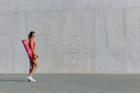 Woman in sportswear walks with a yoga mat and headphones, ready for an outdoor workout.の写真素材