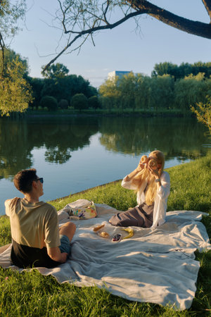 A man and a young woman enjoy food and laughter on a blanket by a sunny pond.の写真素材