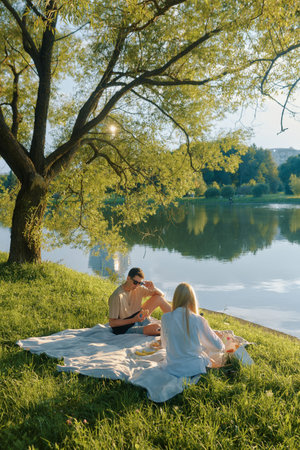 A man and a young woman relax on a blanket by the pond, sharing food and enjoying nature.の写真素材