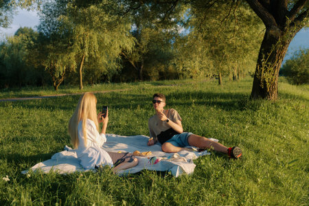 Two friends sit on a blanket under a tree, enjoying food and drinks by a peaceful pond.の写真素材