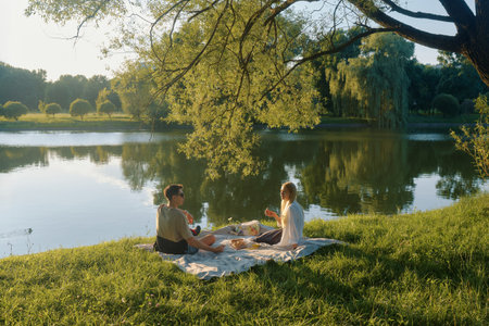 A man and woman picnic together on a blanket beside a calm pond at sunset.の写真素材