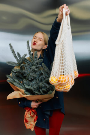 Woman with a playful expression proudly displays a bouquet of evergreens and a net bag of oranges.の写真素材