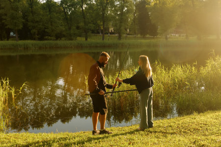 Man and woman are fishing together near a calm pond, enjoying the warm sunlight and nature.の写真素材