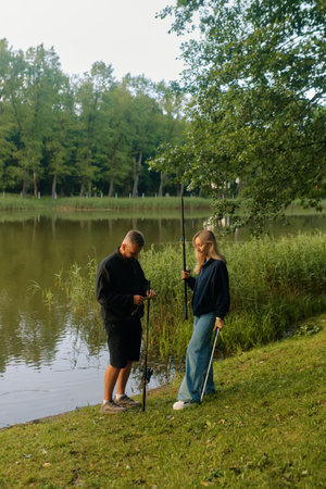 Man and woman prepare fishing rods by the calm pond with greenery all around them.の写真素材