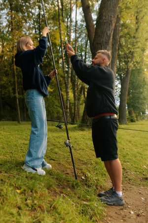 Man helps woman prepare fishing rod near a tranquil pond as sunlight filters through trees.の写真素材