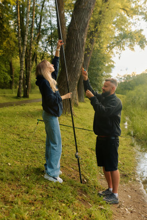 A man assists a woman as they prepare fishing gear by a tranquil pond surrounded by trees.の写真素材