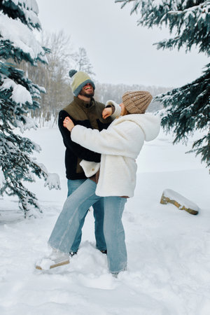 A couple joyfully embraces in a snowy setting, surrounded by tall evergreen trees. Their playful interaction highlights the beauty of winter.の写真素材