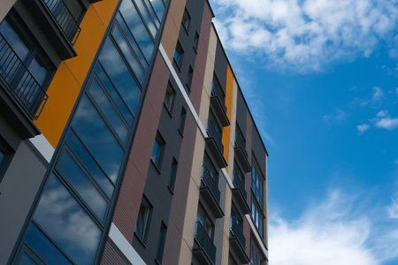 A low-angle view of a modern apartment building with large windows and balconies. The building is painted in shades of gray, brown, and beige, with an accent of bright orange.の写真素材